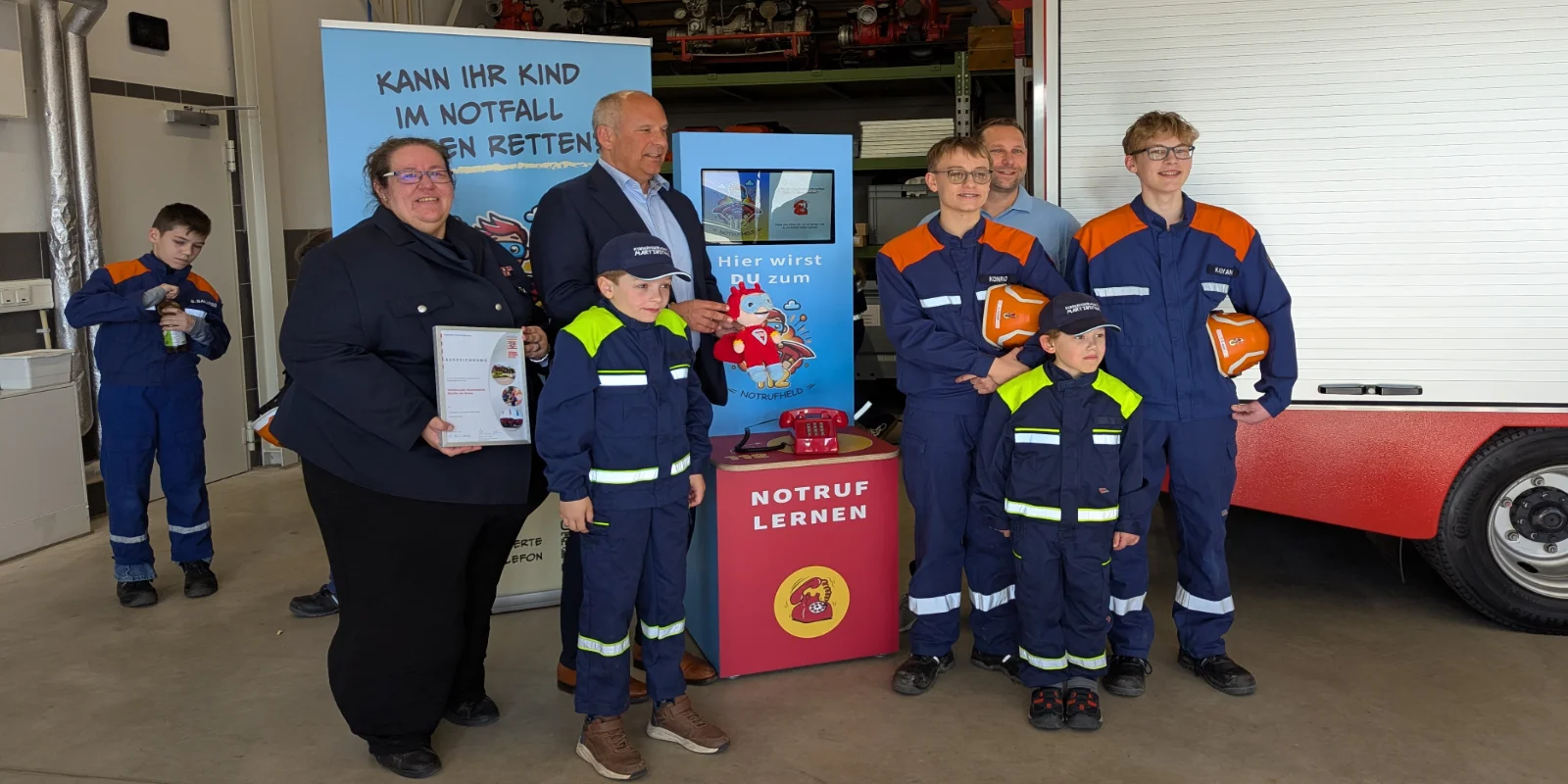 Feuerwehr des Monats Gruppenfoto bei der Auszeichnung zur Feuerwehr des Monats durch den INnenminister mit Kindern und Jugendlichen in Feuerwehrkleidung neben dem interaktiven Lernsystem „Notrufheld“