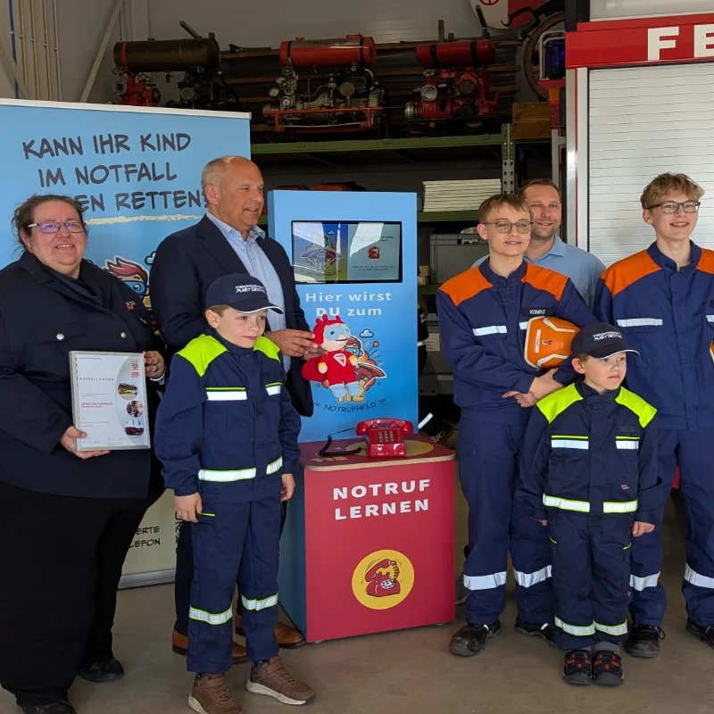 Gruppenfoto bei der Azuszeichnung zur Feuerwehr des Monats durch den INnenminister mit Kindern und Jugendlichen in Feuerwehrkleidung neben dem interaktiven Lernsystem „Notrufheld“